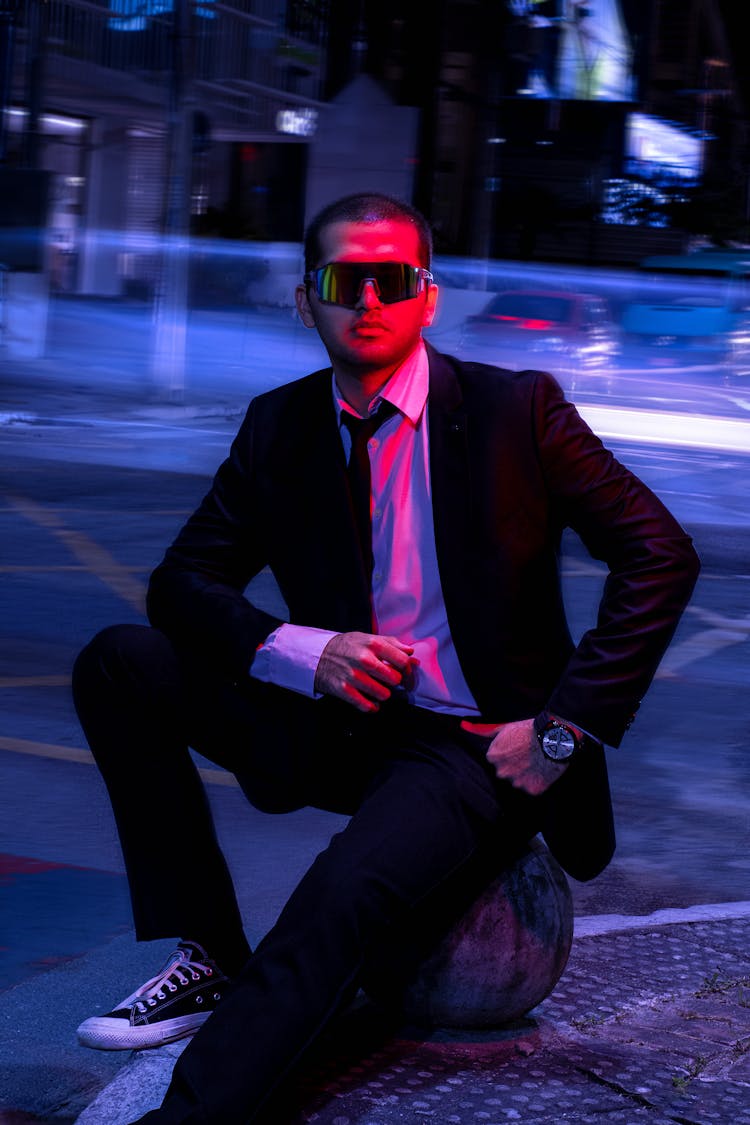 Man In Black Suit Wearing Red Sunglasses Sitting On A Cobble Stone