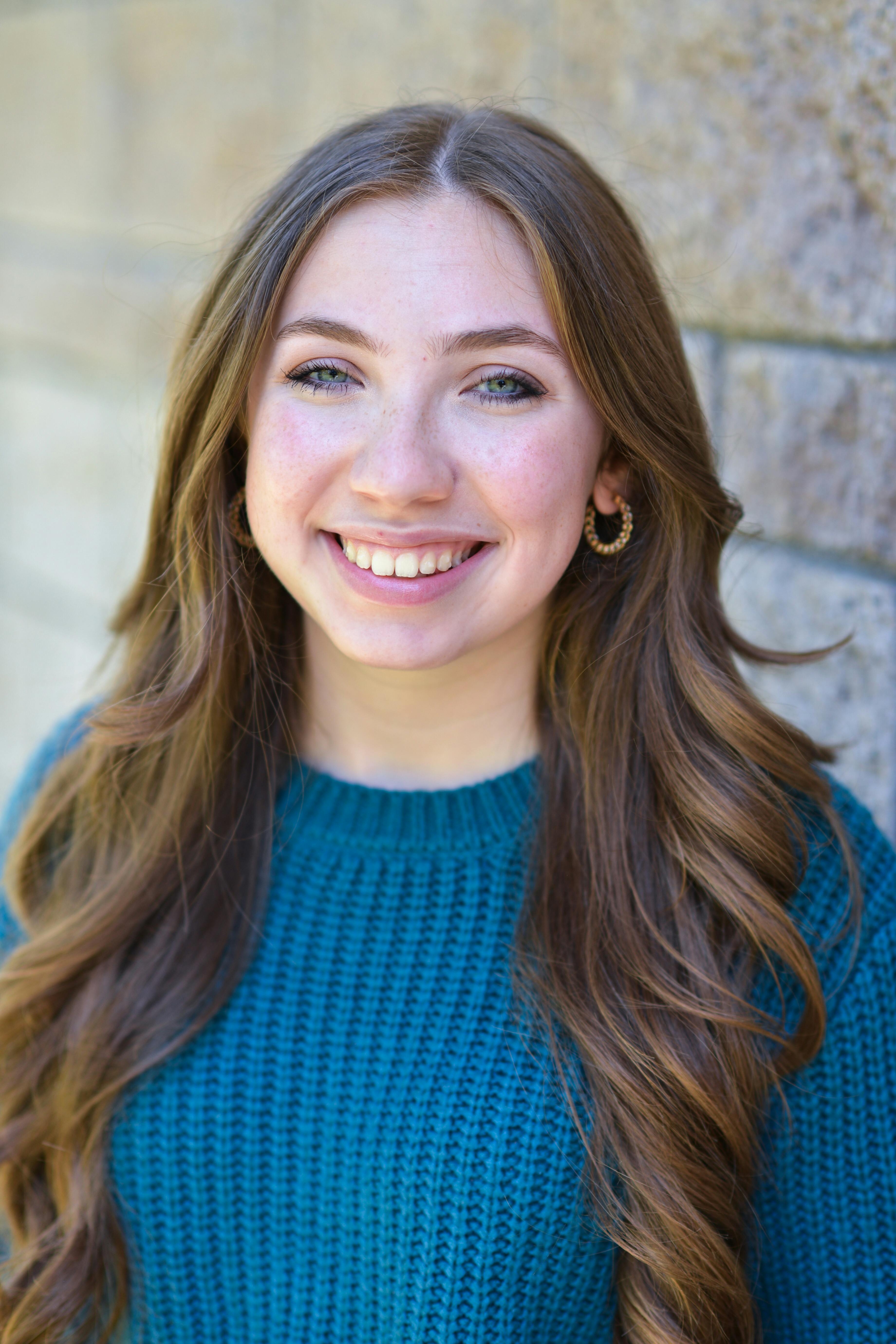 Close-up portrait of a smiling woman in a blue knitted sweater, standing against a brick wall.