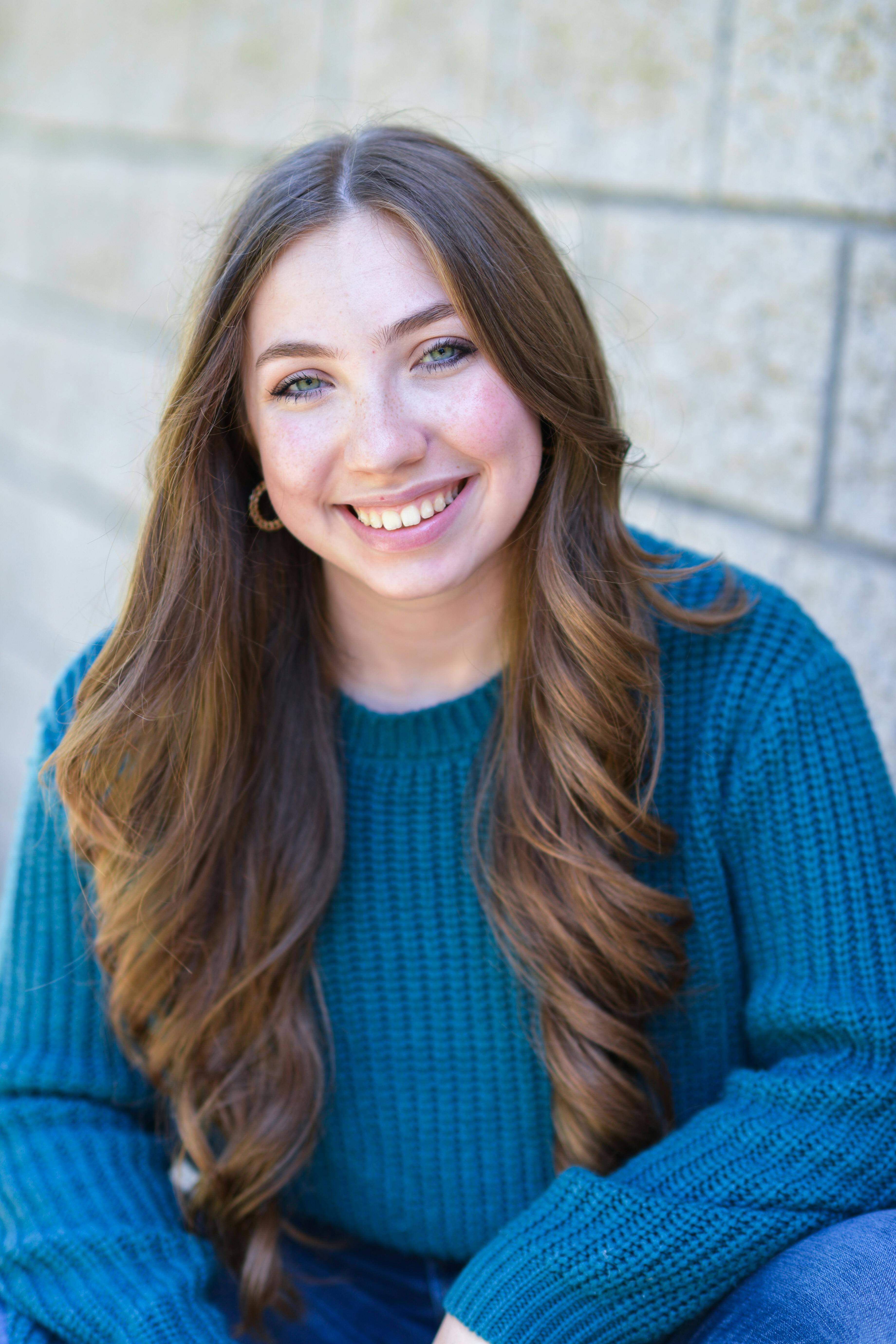A cheerful woman in a teal sweater sits against a brick wall, beaming with joy. Captured outdoors, showcasing natural beauty.