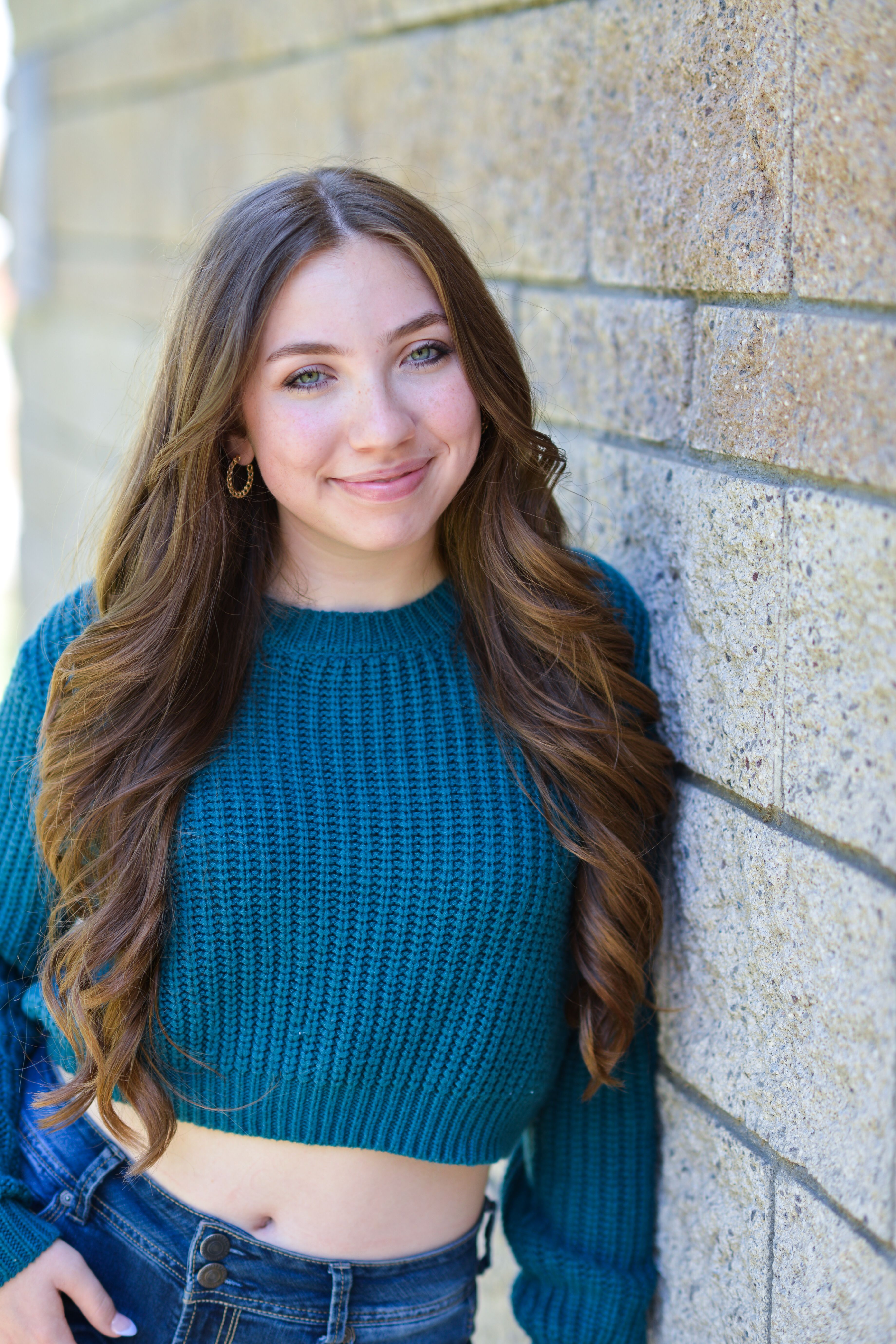 Smiling young woman in a teal knitted sweater leaning against a wall, outdoors.