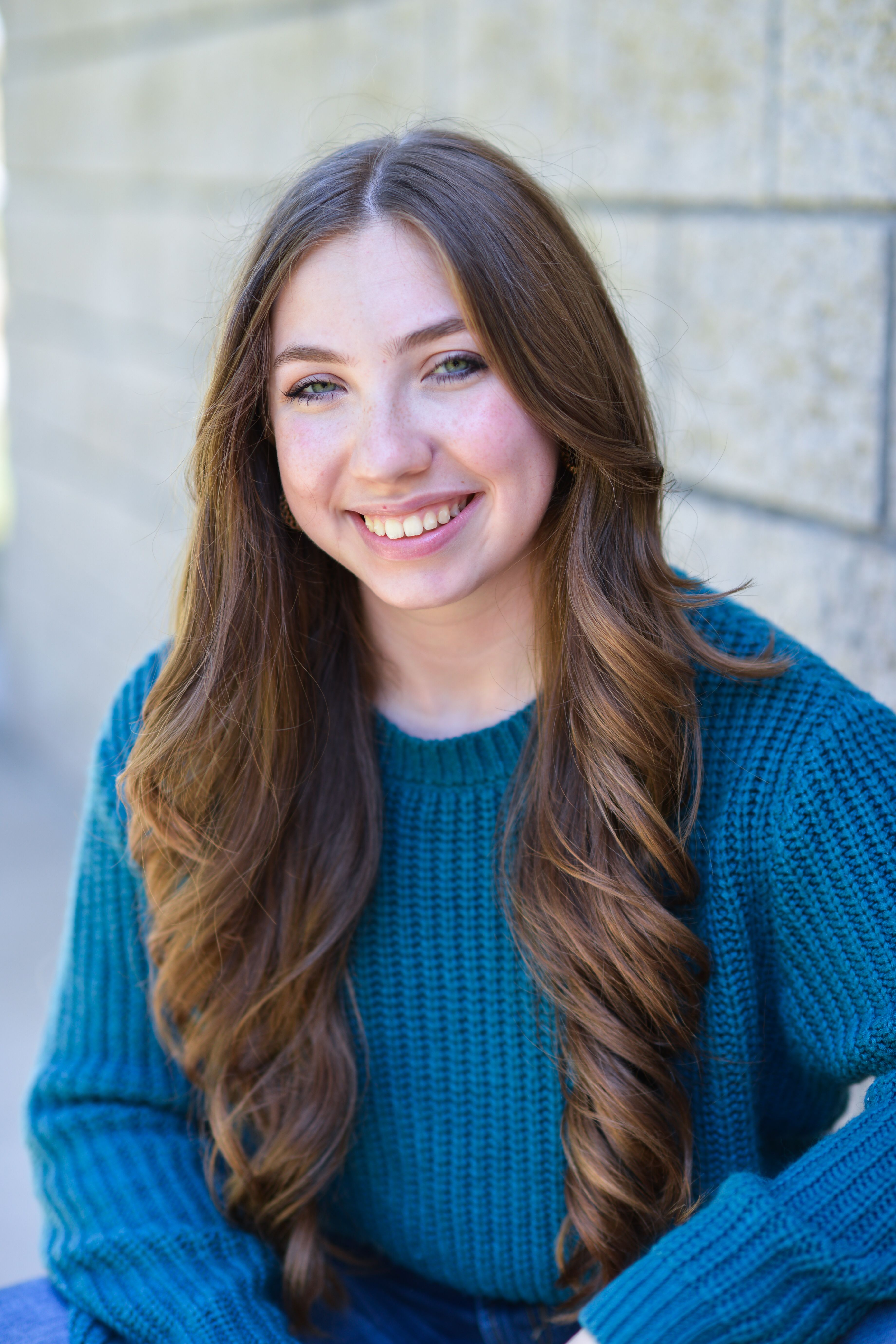 Portrait of a smiling woman in a teal knit sweater sitting outdoors by a stone wall.
