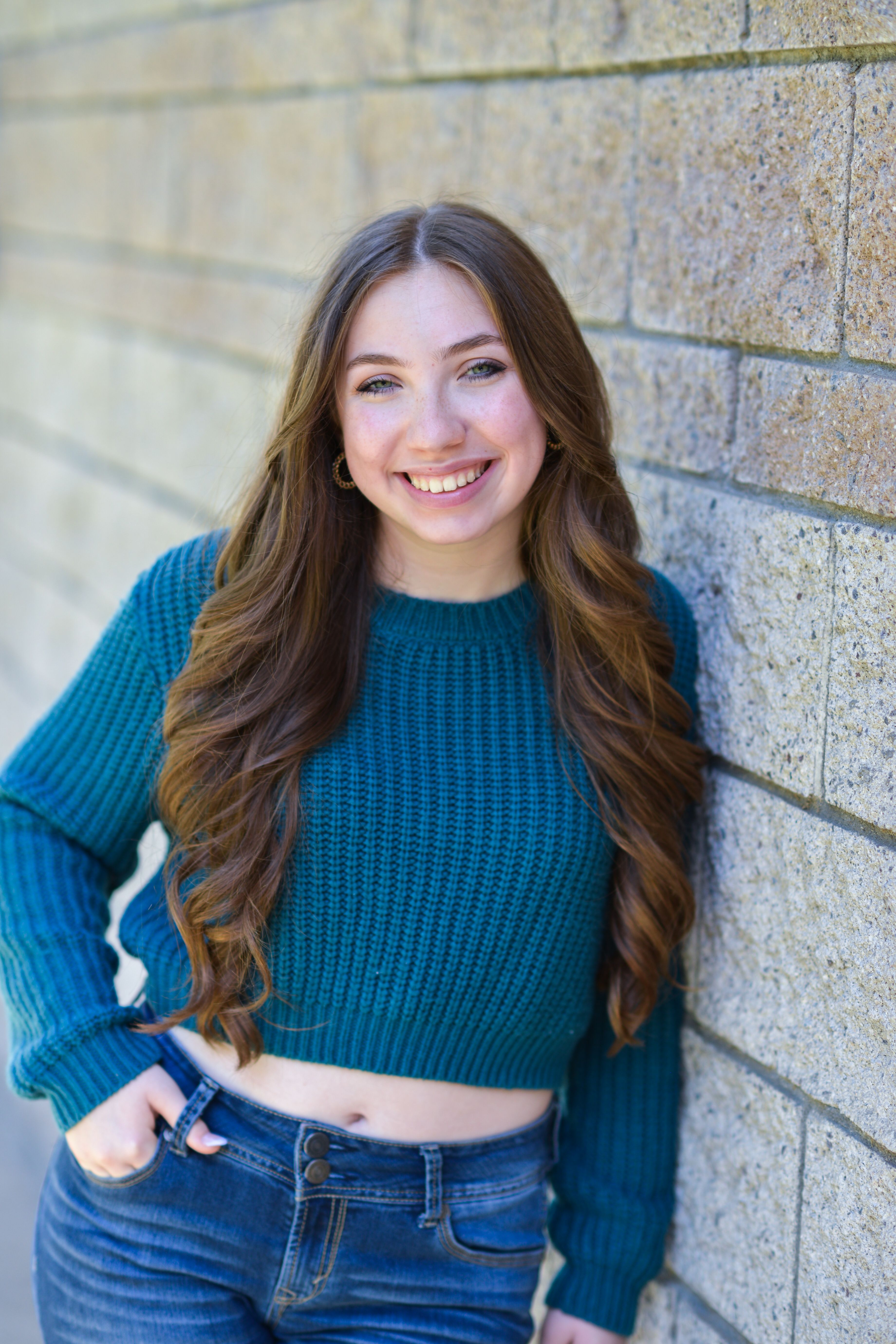 A youthful woman in a teal sweater poses against a brick wall, exuding warmth and friendliness.