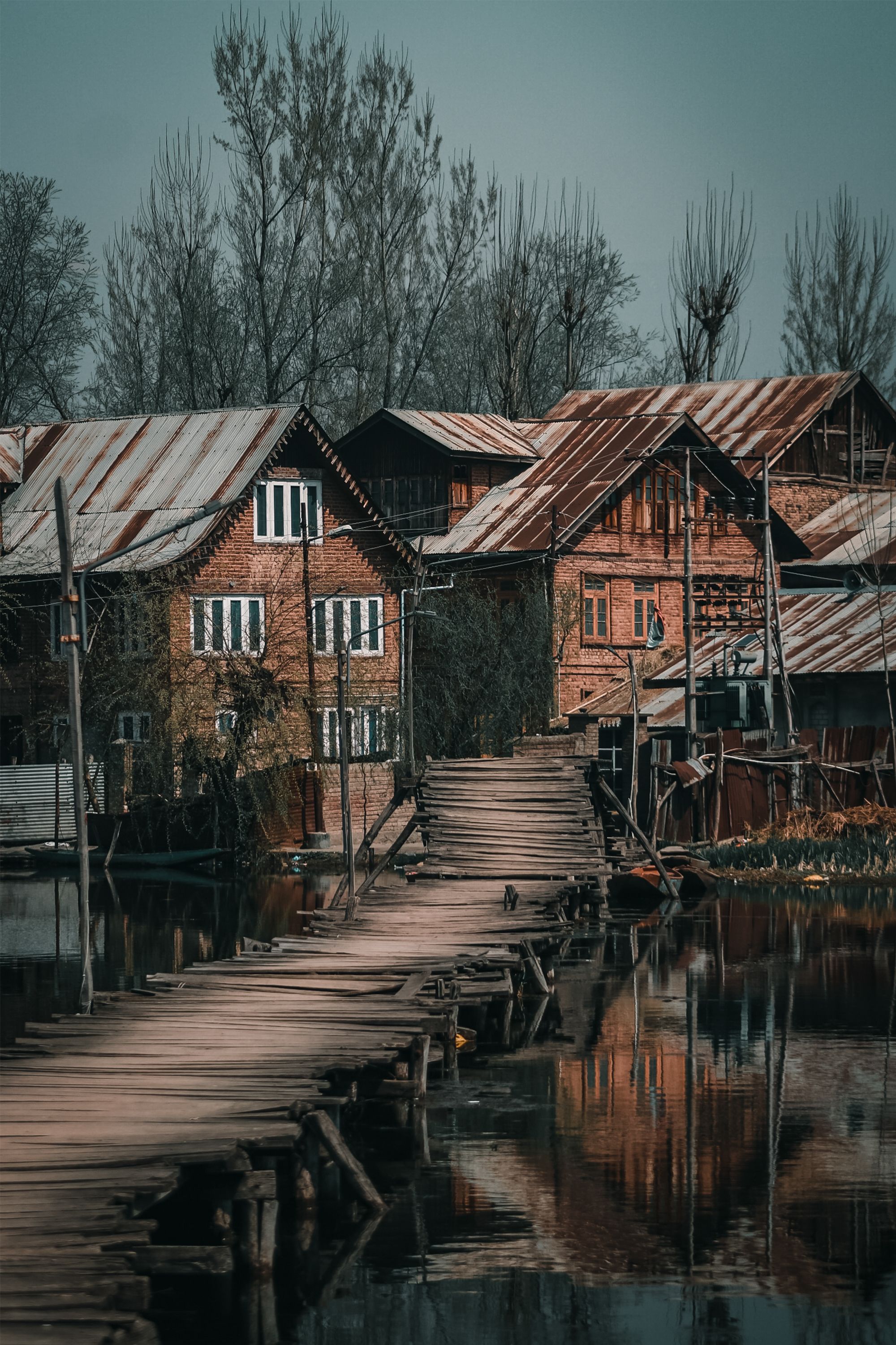 A rustic wooden footbridge leads to traditional houses in a serene village in Srinagar.