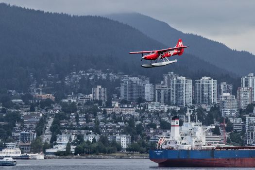 A striking red seaplane soars above a bustling waterfront cityscape with a backdrop of high-rise buildings.