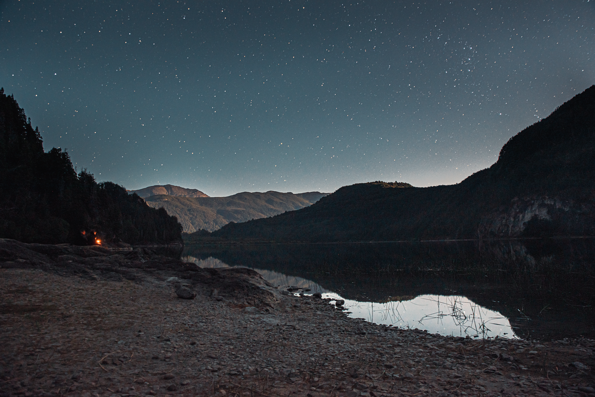 A starry night sky over a still Patagonian lake, mountains silhouetted, a small campfire glowing on the shore