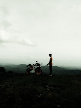 Silhouetted biker standing with motorbike against a mountainous backdrop in India, capturing adventure and serenity.