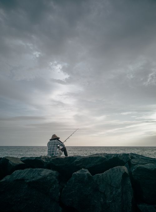 Free A lone fisherman sits on rocks by the ocean under a cloudy sky in Kozhikode, India. Stock Photo