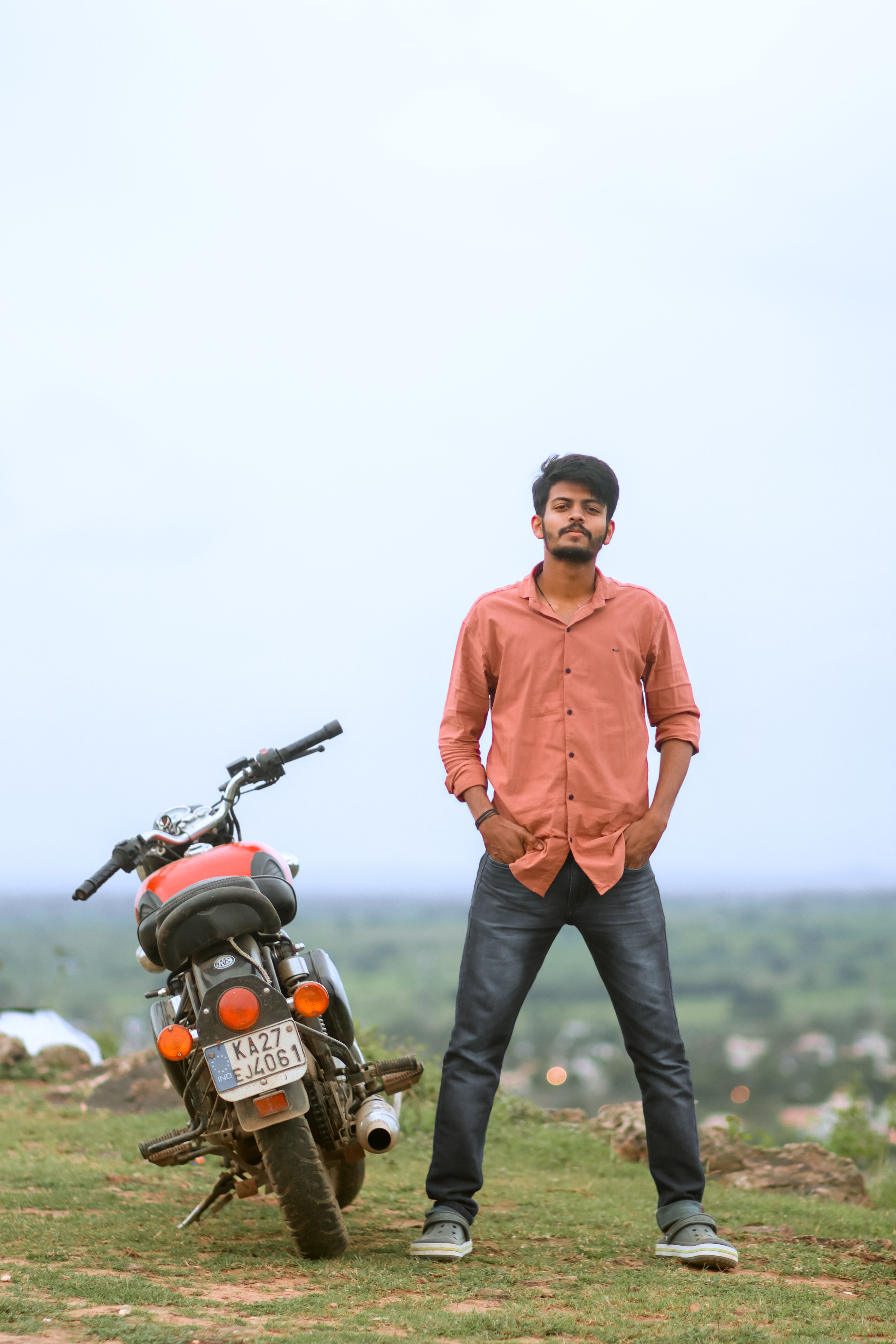 Young man stands confidently beside motorcycle on a hill with scenic background.