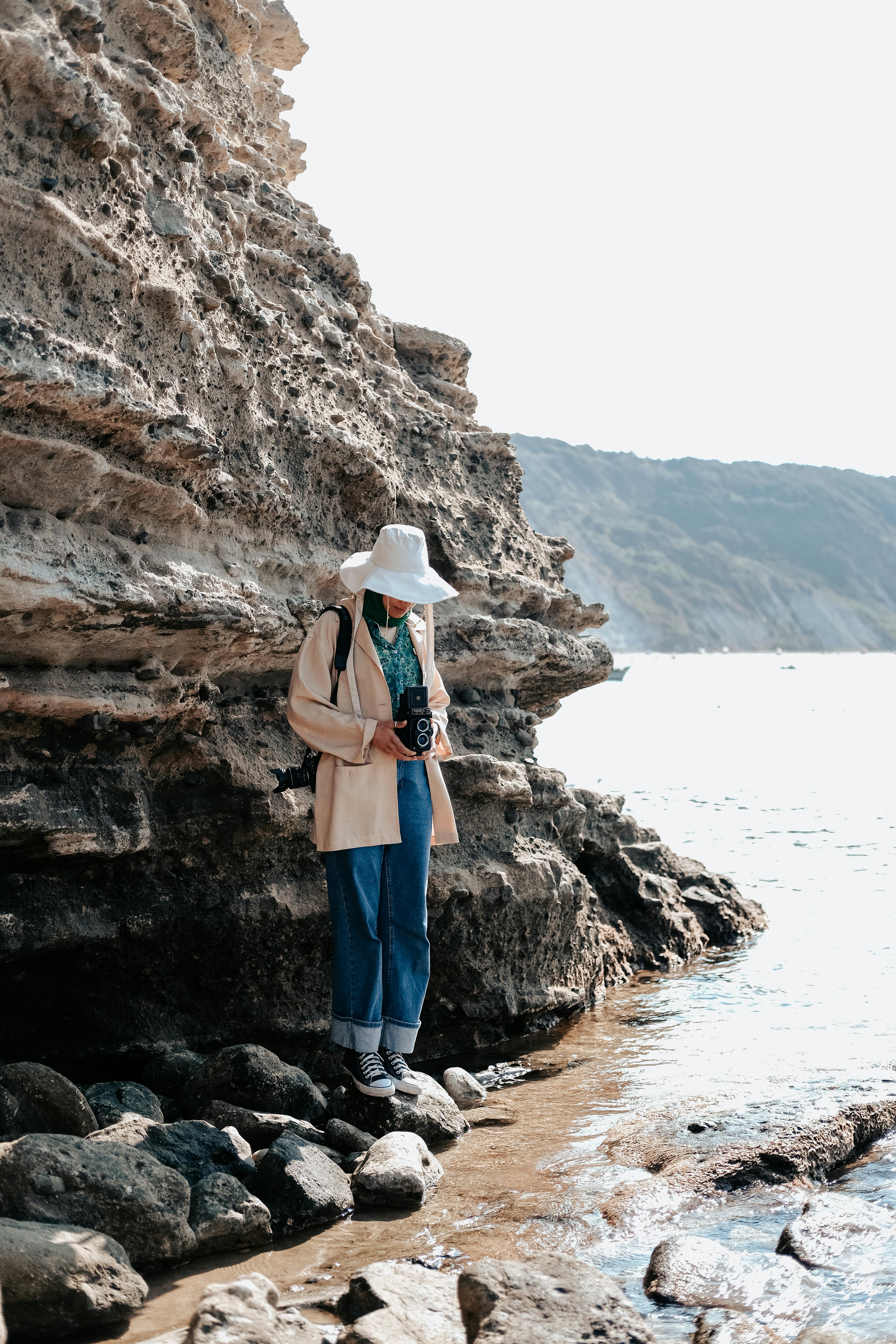 A woman in a wide-brimmed hat photographs the seashore beside a rocky cliff on a sunny day.
