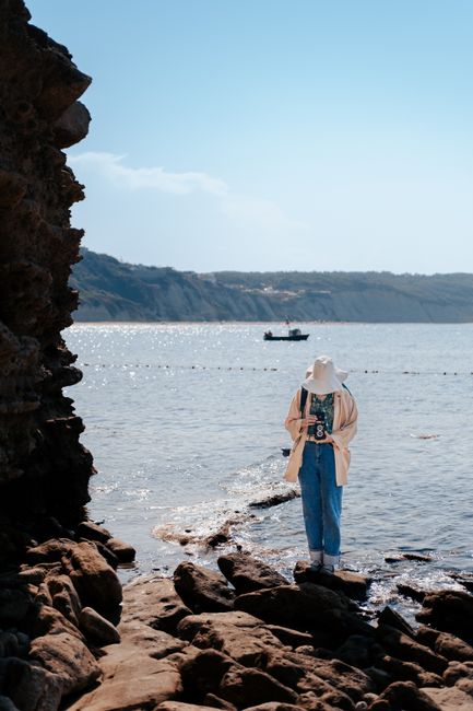 A woman in casual attire enjoys a sunny day by the sea near a rocky cliff and boat.