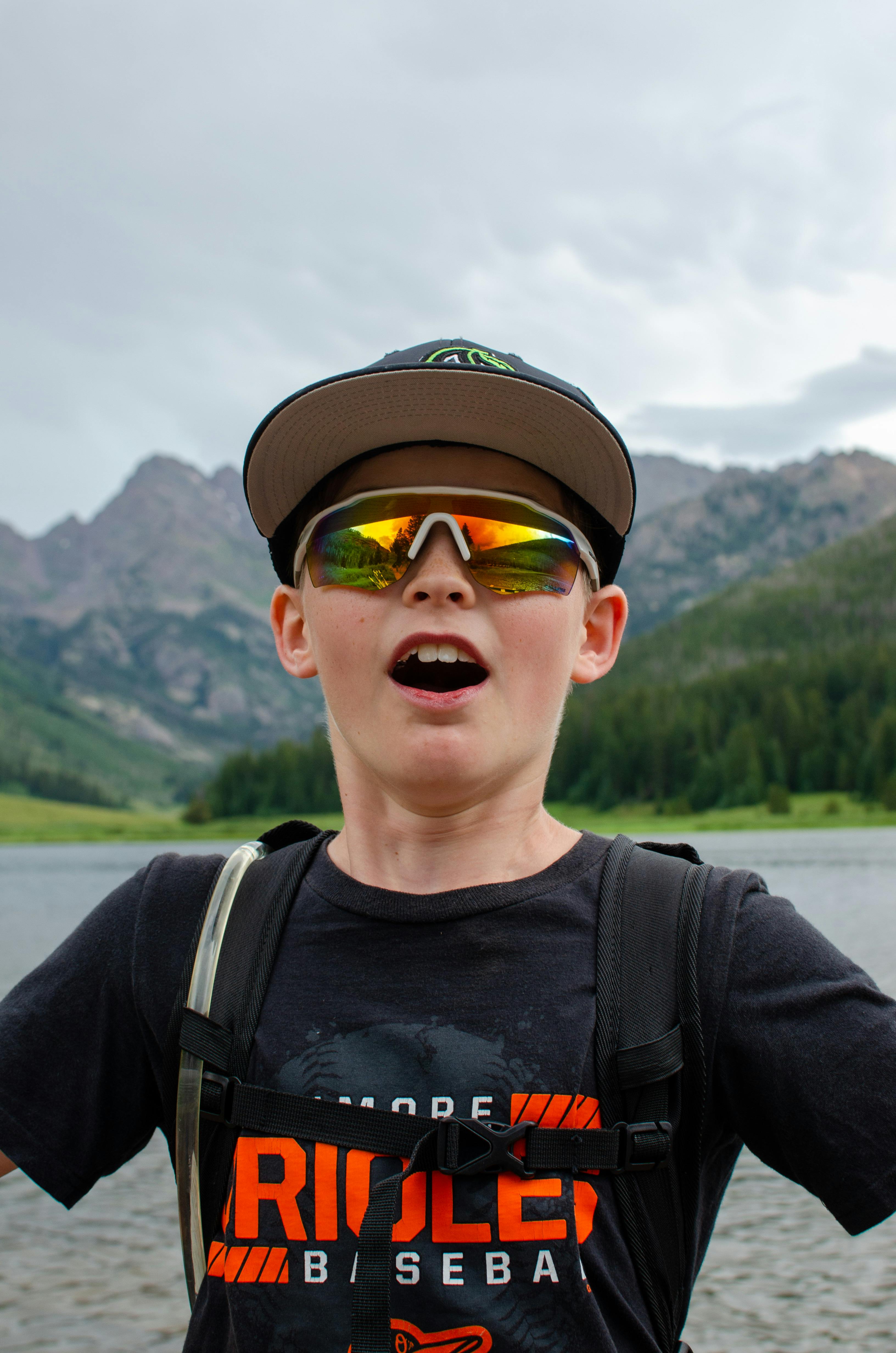A boy wearing sunglasses and a backpack enjoys a day outdoors by a mountain lake.