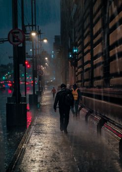 People walking on wet city sidewalk at night under streetlights in the rain.