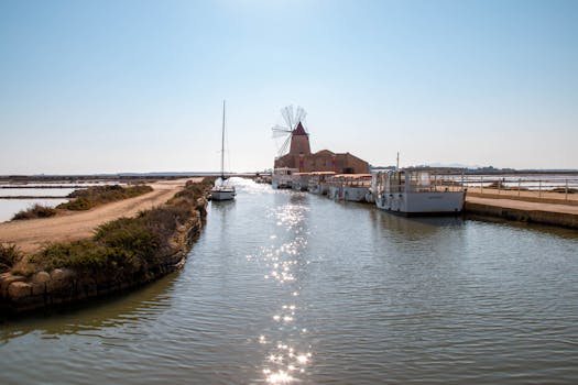 Scenic view of windmill and boats on a canal in Marsala, Sicily, Italy under clear blue sky.