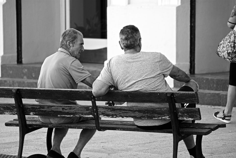 Dos hombres mayores sentados en un banco, capturados en una interacción espontánea en blanco y negro.