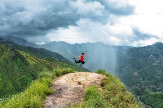 Man in red shirt jumps energetically on a mountain cliff with a dramatic cloudy sky backdrop.
