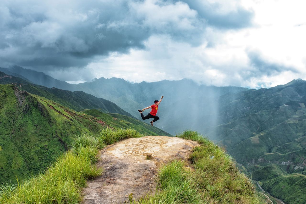 man in red shirt jumps energetically on a mountain cliff with a dramat