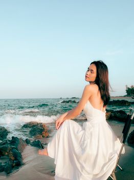 A woman in a white dress sits on the rocks by the ocean, enjoying a peaceful sunset moment.