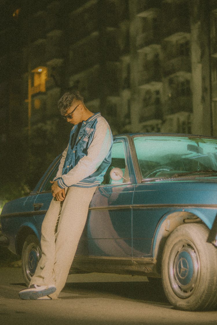 Man Wearing A Varsity Jacket Leaning On A Blue Car