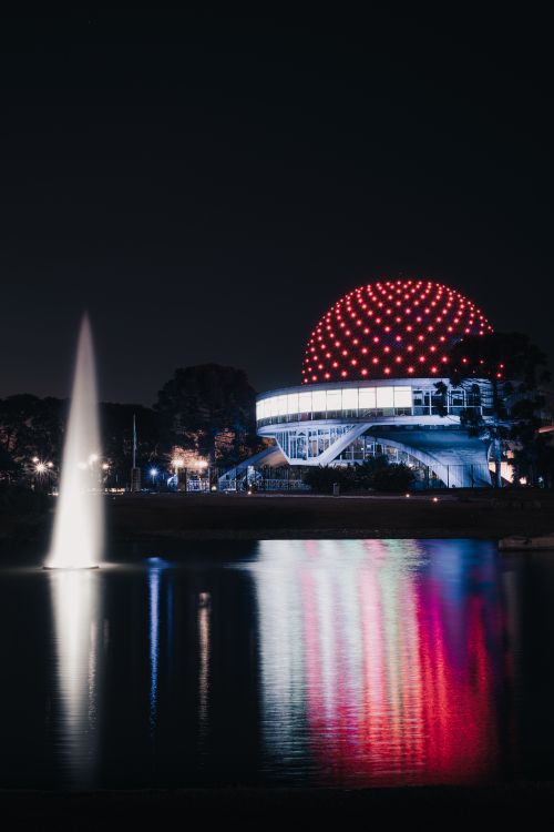 Free Majestic night view of a red-lit planetarium with reflection on water and a fountain display. Stock Photo