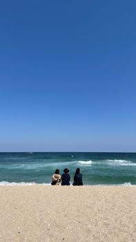 Three people sit together on a sunny beach in Yangyang, Korea, looking at the sea under a clear blue sky.