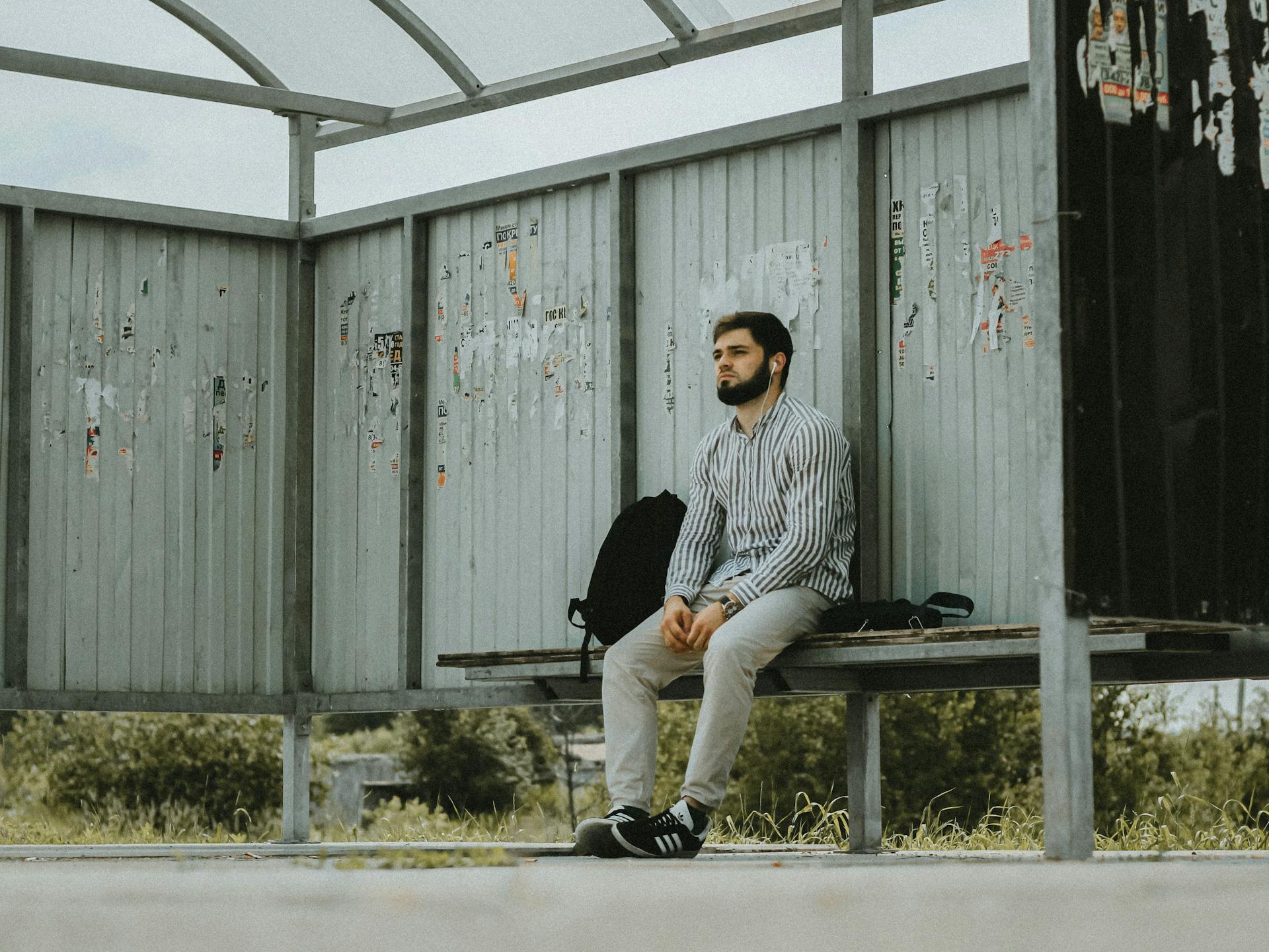 A young bearded man sits at an outdoor bus shelter, waiting patiently on a sunny day.