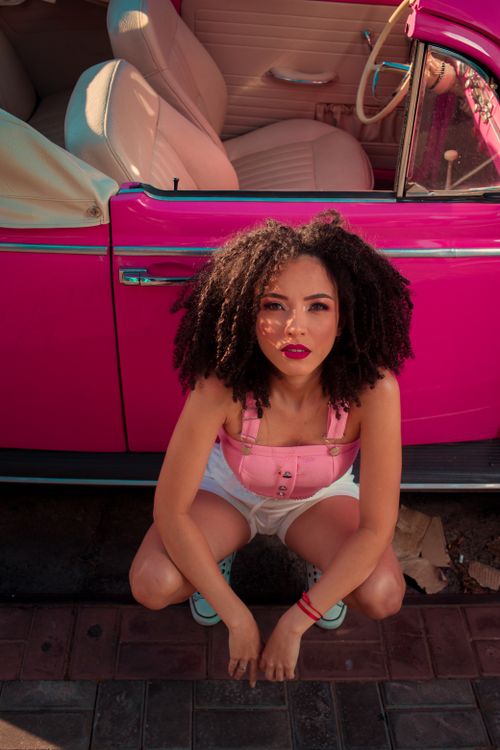 Free An adult woman with curly hair squats by a pink vintage car, creating a vibrant and stylish scene. Stock Photo