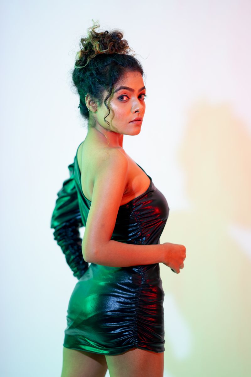 A fashionable woman in a shiny black dress posing indoors with colorful lighting.