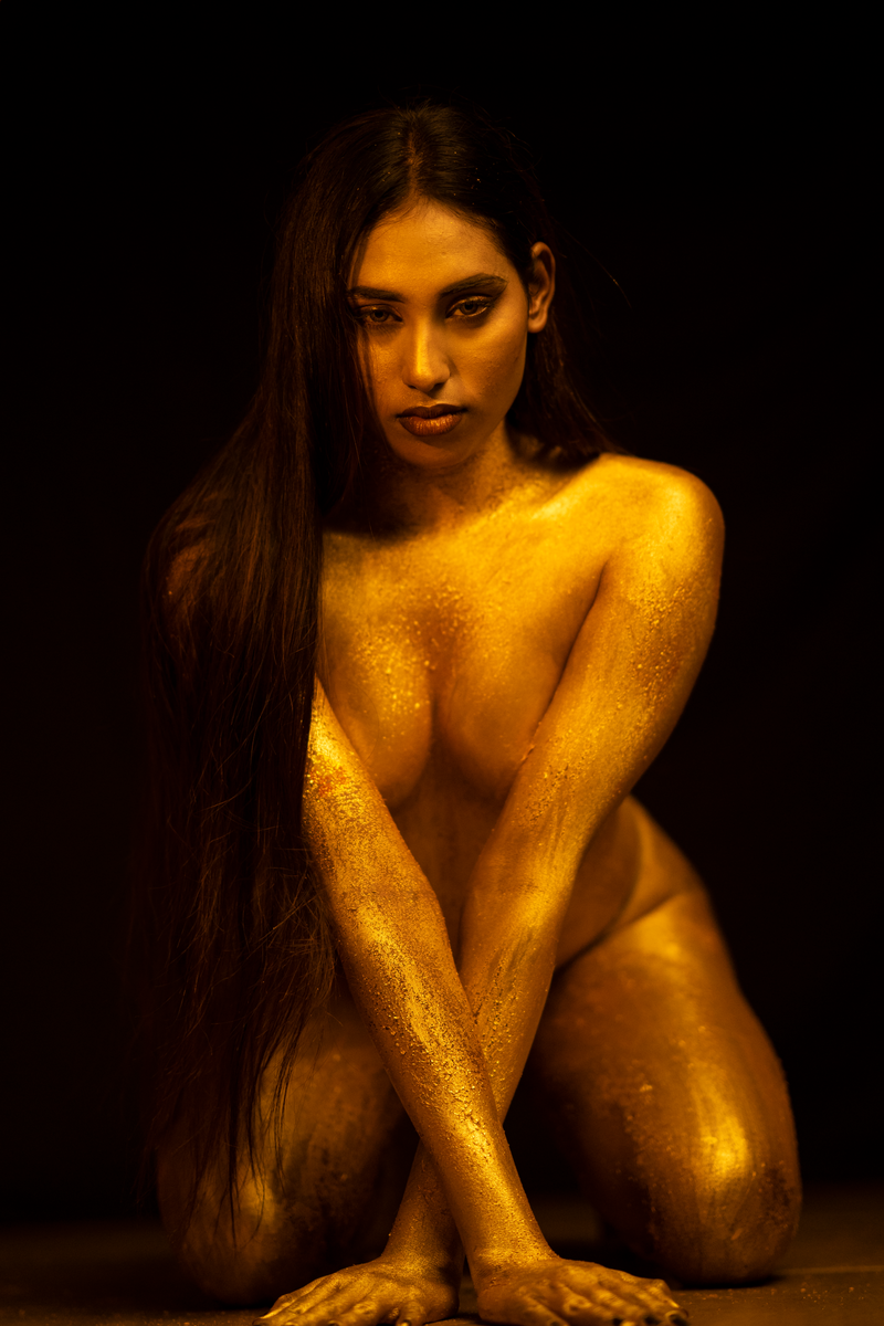 Striking studio portrait of a woman covered in gold paint against a dark background.