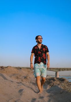 Young man in fashionable summer attire strolling on a beach with a clear blue sky backdrop.