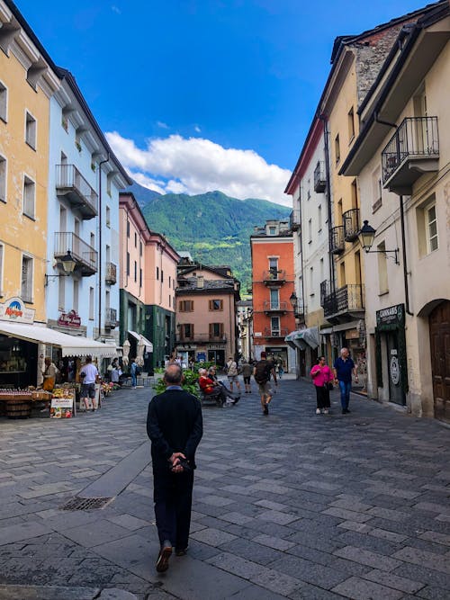 An Old Person Walking on Alley Between Buildings Near a Mountain Under Blue Sky