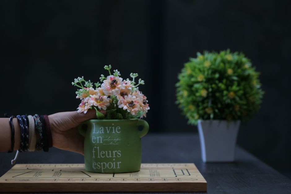 A hand arranging flowers in a green pot with text indoors on a wooden table.
