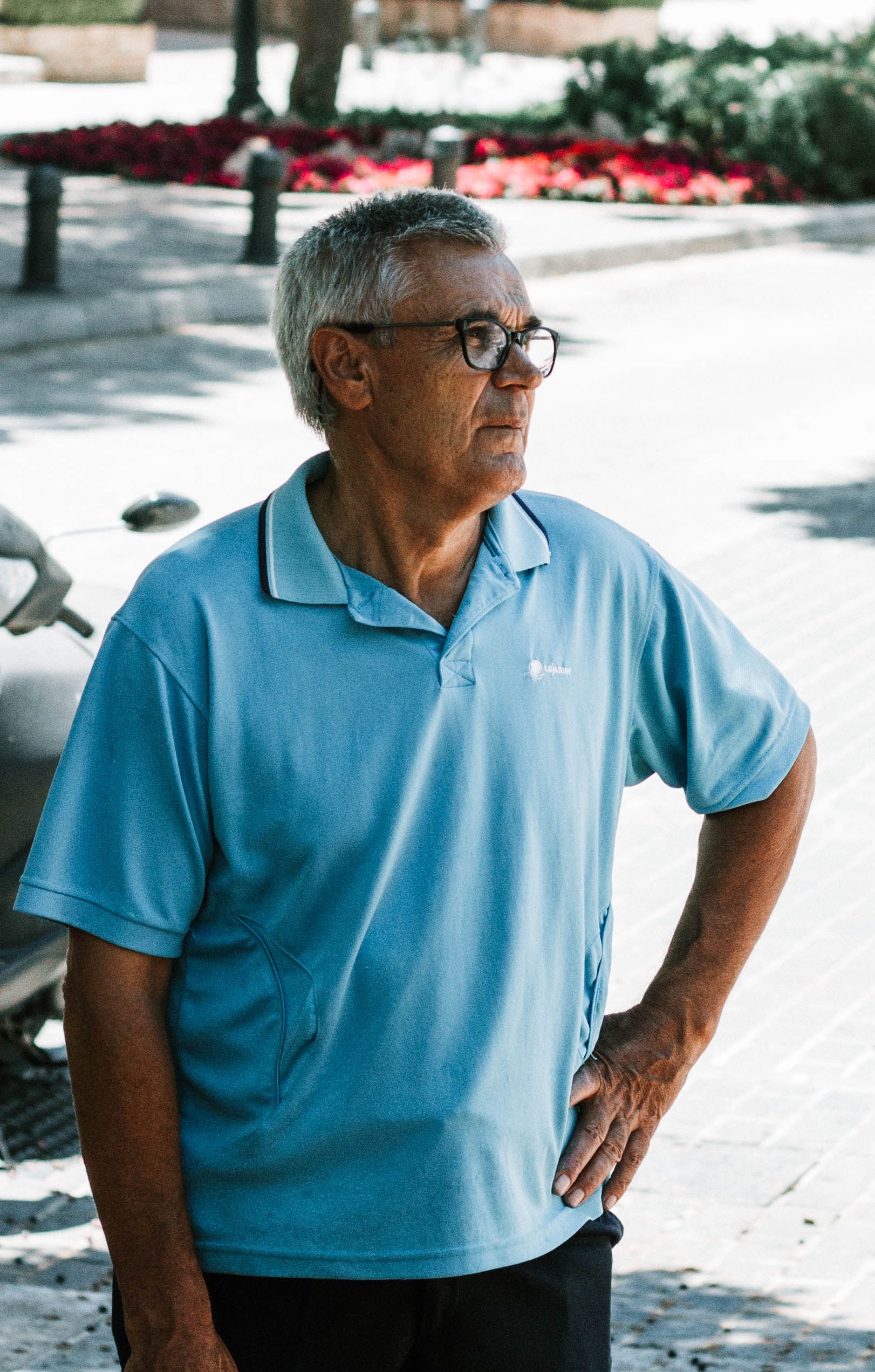 An elderly man with glasses stands contemplatively outdoors in a relaxed pose.