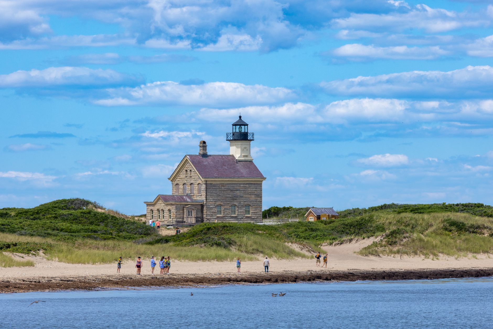 A serene view of Block Island Southeast Lighthouse with people enjoying the beach under a blue sky.