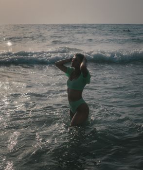 A woman in a bikini poses in the ocean during sunset, embracing a serene seaside vibe.