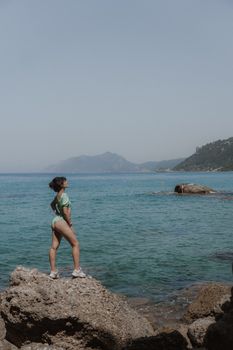 A woman wearing a swimsuit stands on rocks by the sea on a clear day, gazing at the horizon.