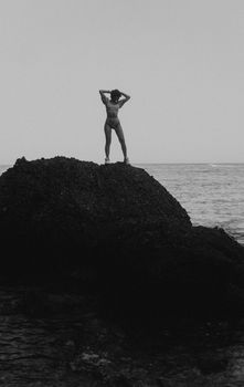 A lone woman standing on a rock by the sea, captured in grayscale.