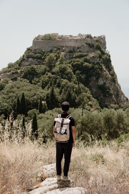 A lone backpacker stands facing an ancient, tree-covered mountain fortress under a bright sky.