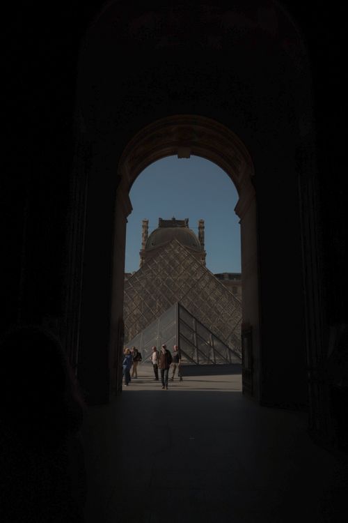 Free Tourists walk towards the iconic Louvre Pyramid in Paris through a shadowed archway. Stock Photo