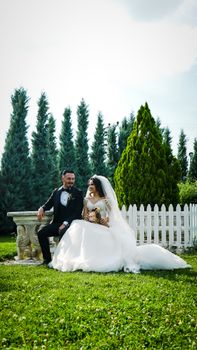 Stunning outdoor portrait of a newlywed couple seated in front of a white fence, bride in gown, groom in tuxedo.