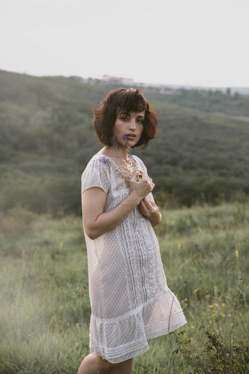 Woman in a White Summer Dress Standing on a Field and Holding Flowers 