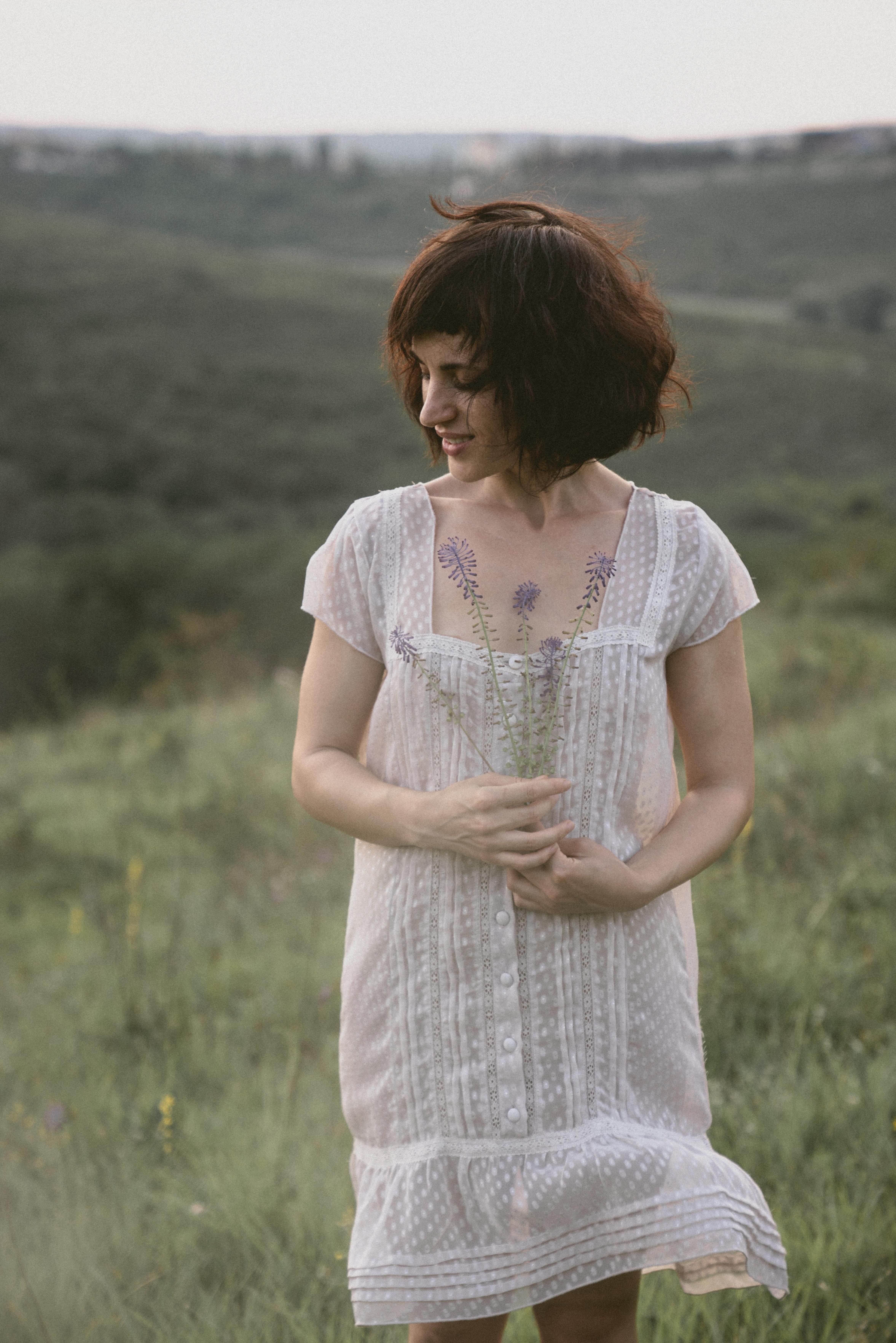 A woman in a white dress holding lavender, standing in a grassy countryside with a serene expression.