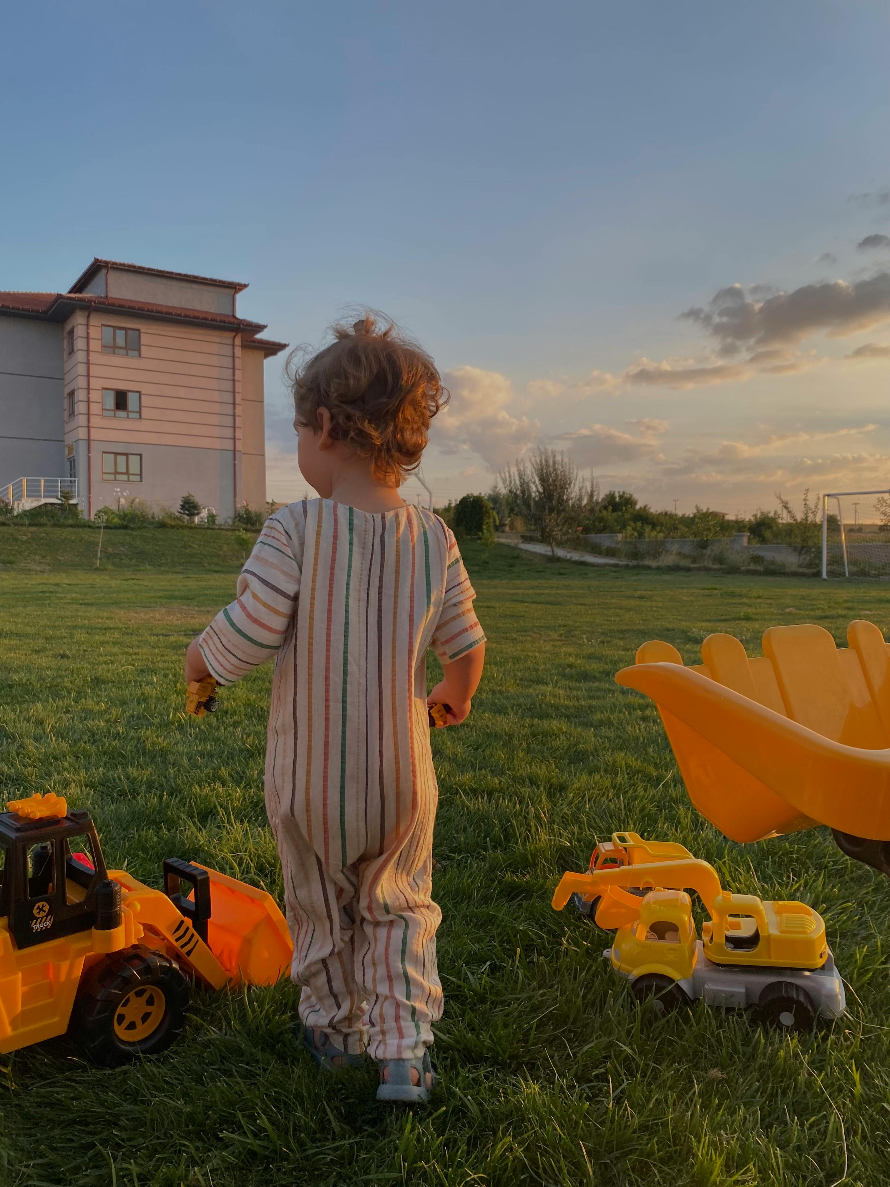 A toddler in pajamas playing with toy trucks on a grassy lawn during sunrise, near a house.