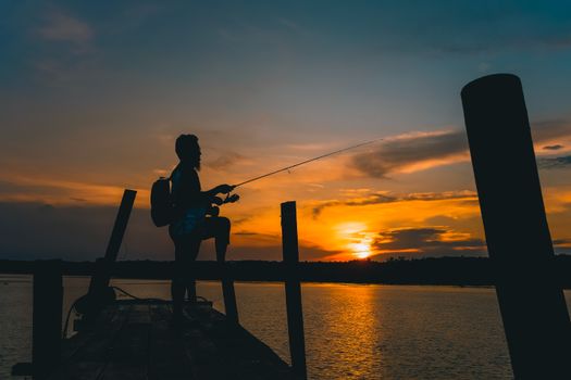 Peaceful silhouette of a fisherman standing on a dock, casting a line at sunset over a serene lake.