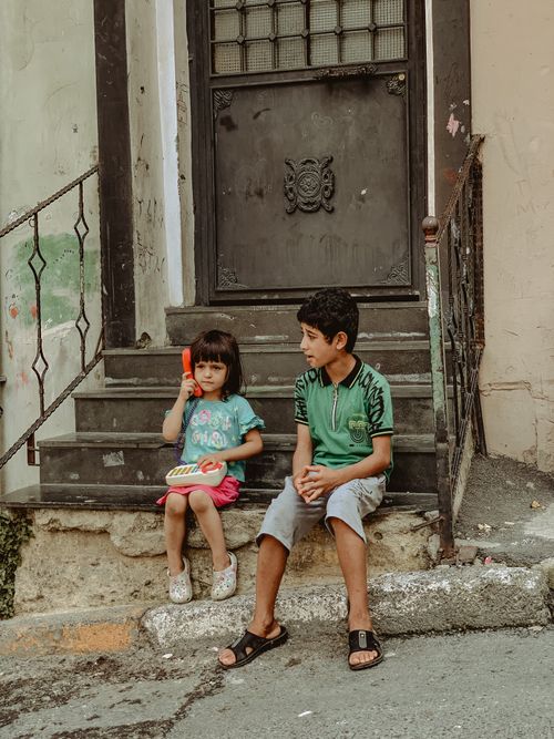 Free Two children sit on steps outside a door, one holding a toy, in an urban street setting. Stock Photo