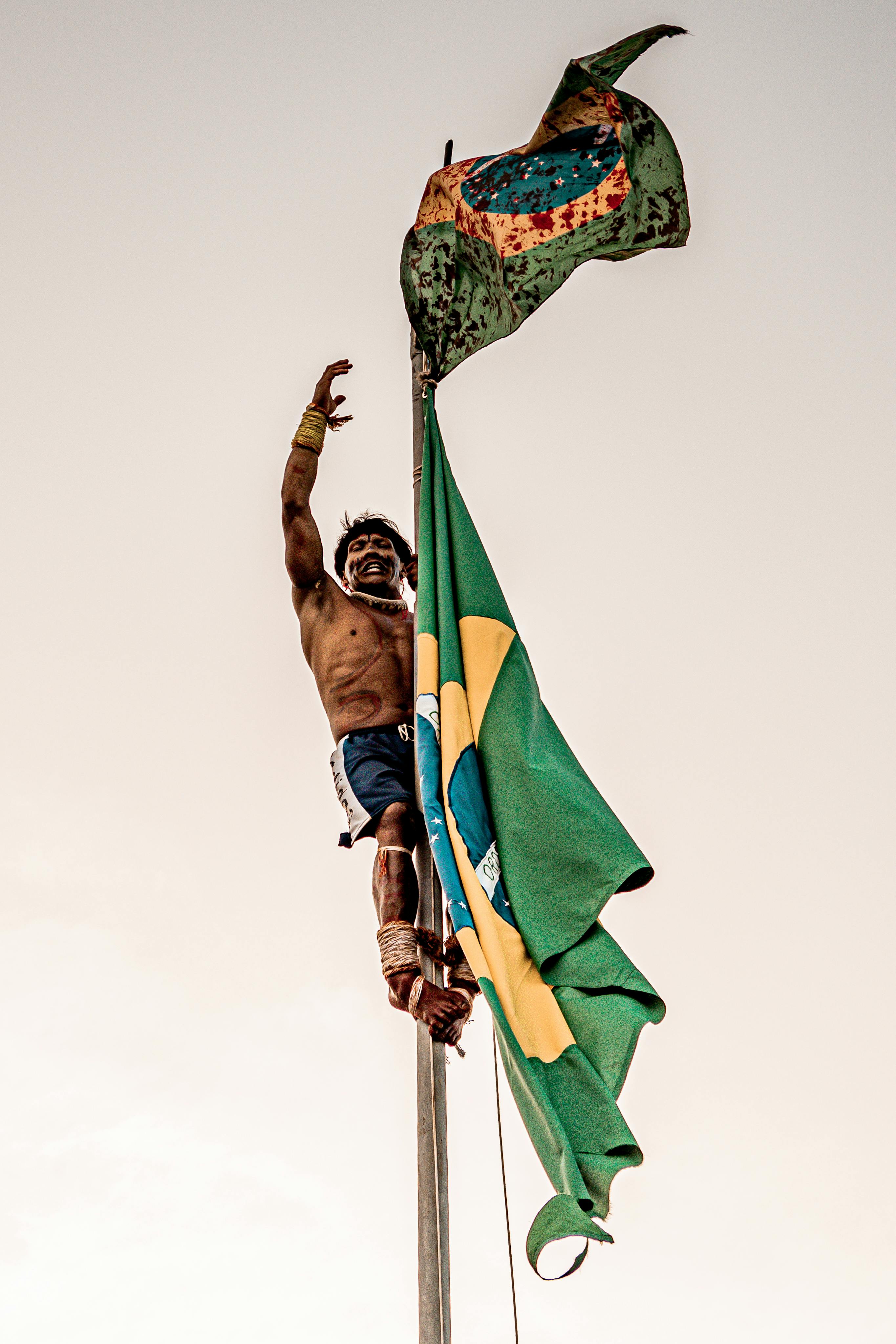 An indigenous man climbs a flagpole holding the Brazilian flag, celebrating cultural pride.