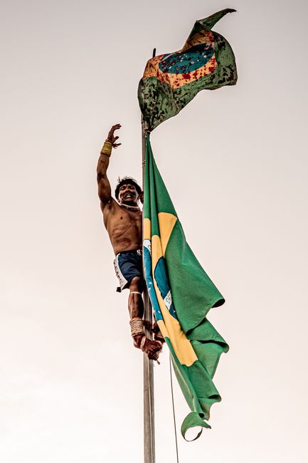 Fotomural Un hombre indígena trepa un mástil sujetando la bandera de Brasil, celebrando el orgullo cultural.