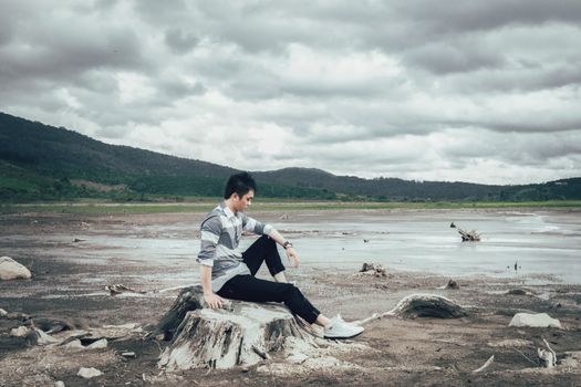 Asian man sitting on tree stump in vast, barren outdoor landscape on a cloudy day.