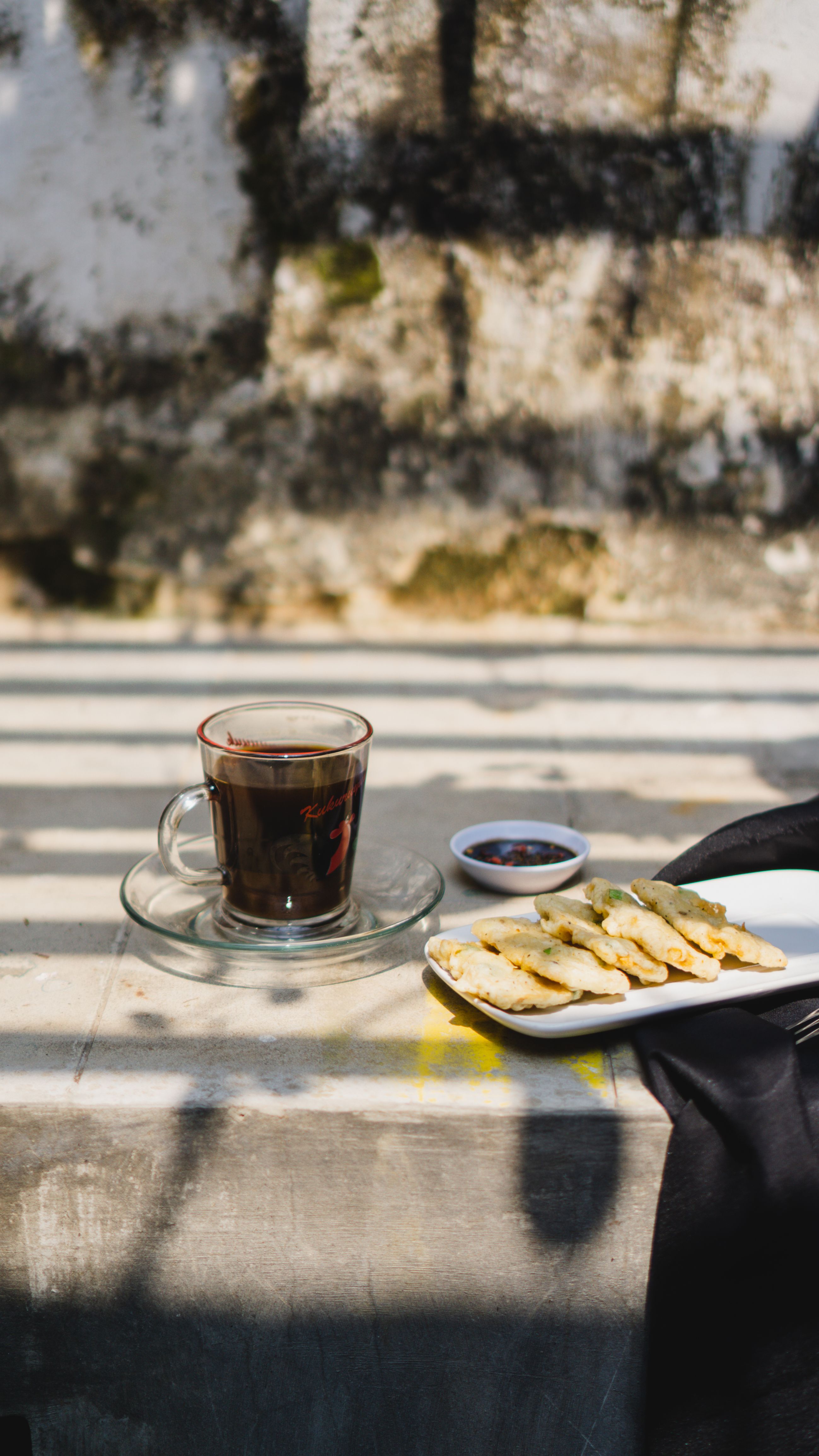 A cup of coffee with snacks arranged on a rustic outdoor table under striped shadows.