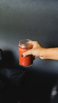 A person holding a clear glass filled with red juice against a neutral background.