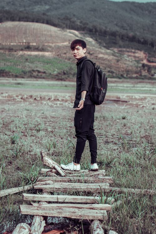 Free Young Asian man with backpack stands in a vast grassy field, surrounded by hills and nature. Stock Photo